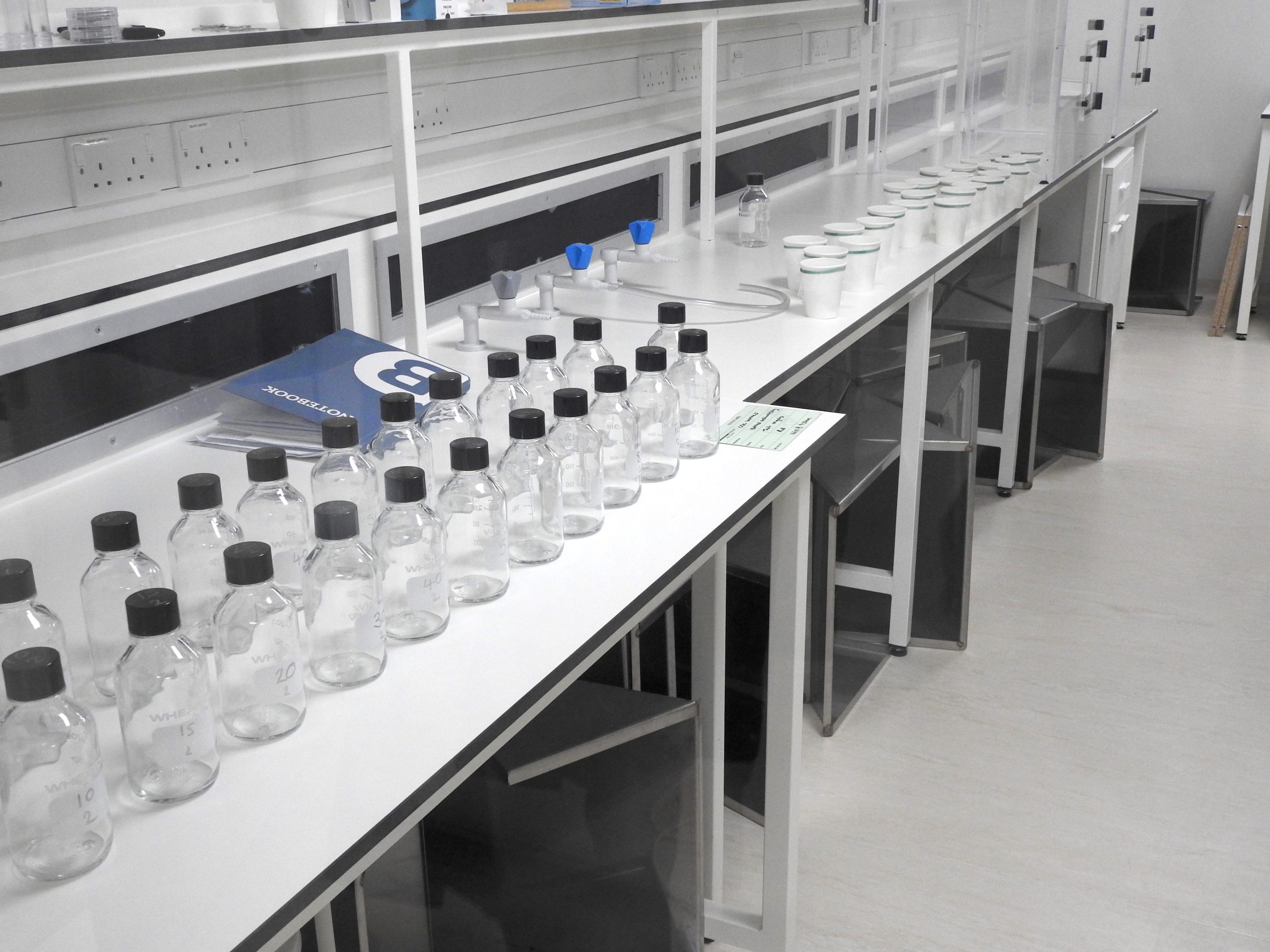 Testing lab. LITE, 2023 White lab bench diagonal from left to right across the image with two long rows of glass bottles with black lids in the forefront of the image and paper cups with netting in the back with a blue lab notebook