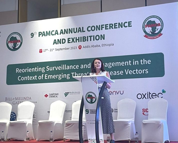 PAMCA 2023. LSTM, 2023 A woman with brown short hair, wearing a white short sleeve shirt and blue trousers standing at a white podium with the PAMCA logo on it on a stage in front of a PAMCA banner and a row of white chairs.