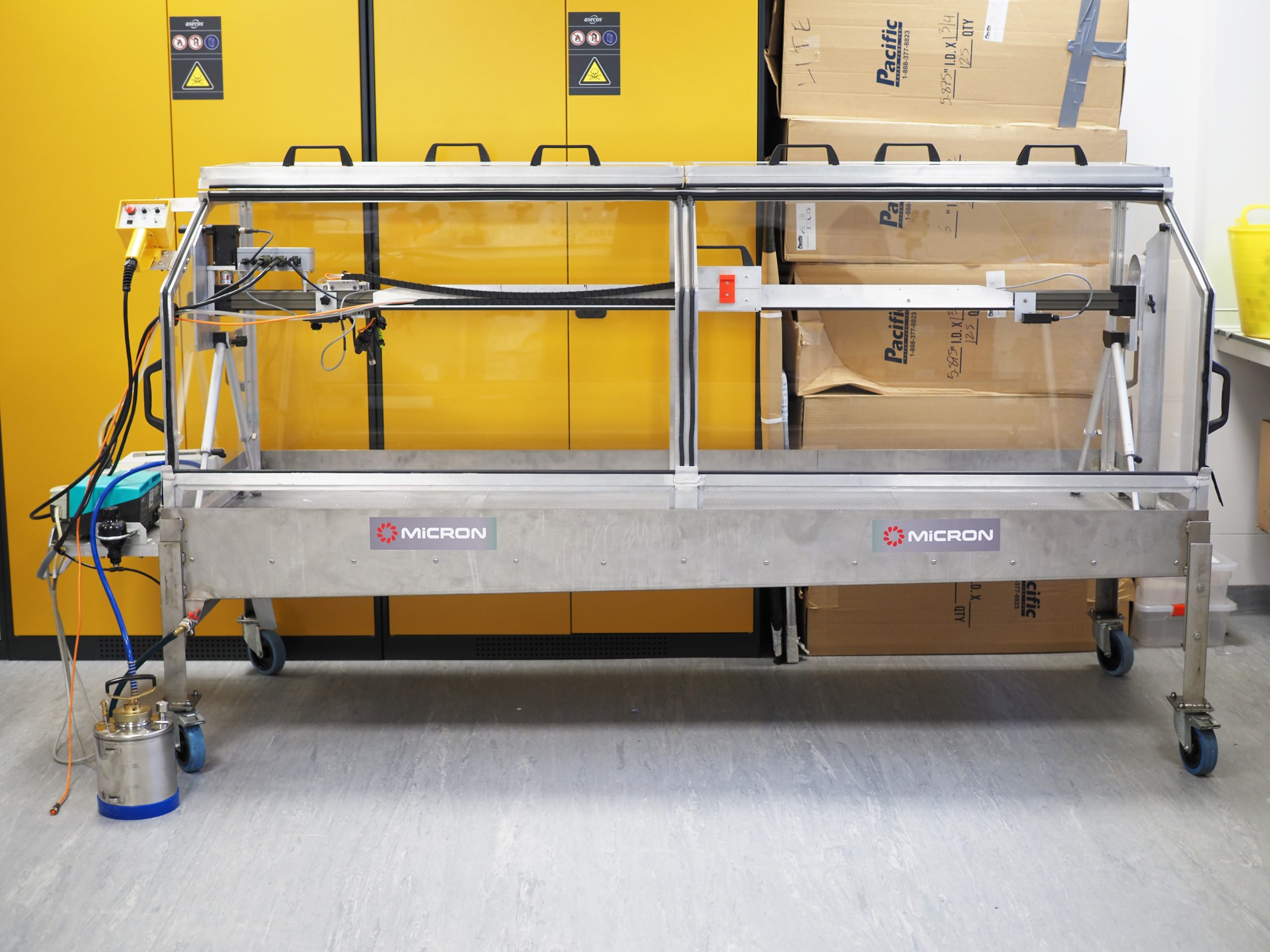 Track Sprayer. I2I. Courtesy of LITE testing facilities. Credit: Kirsten Duda, 2023 Image of the Track Sprayer. a horizontal, rectangular metal structure with glass doors on all sides with a canister at the base on the left side. Bright yellow cabinets are behind the Track Sprayer.